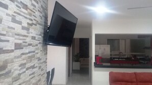 Un salon moderne avec un téléviseur à écran plat fixé sur un mur texturé, un canapé rouge et un coin cuisine visible à travers une porte ouverte.