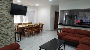 Un salon moderne comprenant un coin repas, un canapé rouge, un téléviseur fixé au mur et une cuisine ouverte sur le salon par une porte sans obstacle.