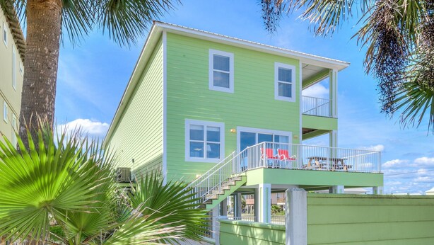 A two-story building with a green exterior, a balcony with red chairs, and a palm tree in the foreground. - Sea Urchins, Unit 2. 5 BR. Beachview. The Beach, so close, for less! Pool. (Gulf Shores)