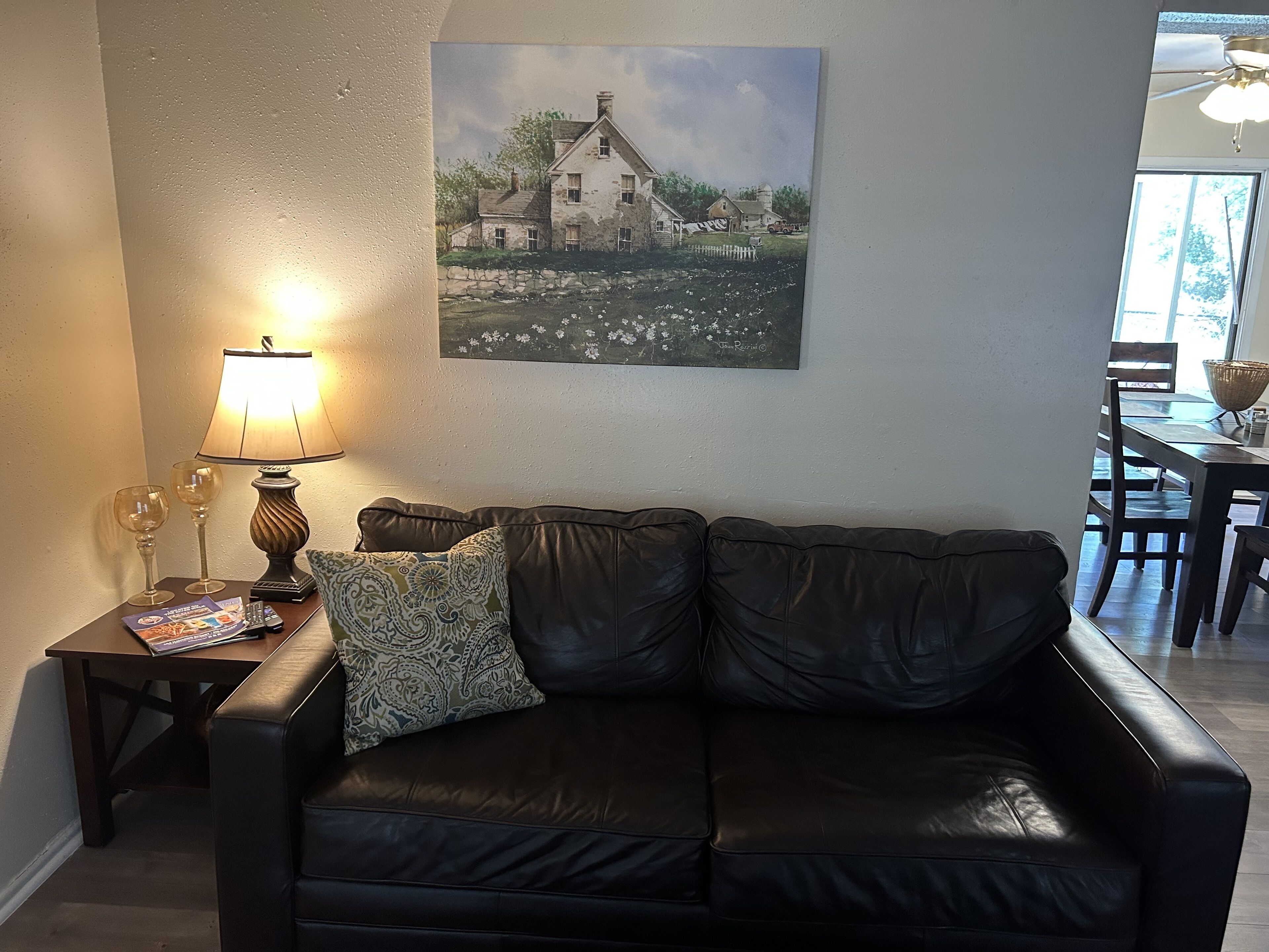 A living room with a black leather sofa, a table with a lamp, two wine glasses, and a framed picture on the wall.