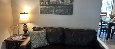 A living room with a black leather sofa, a table with a lamp, two wine glasses, and a framed picture on the wall.