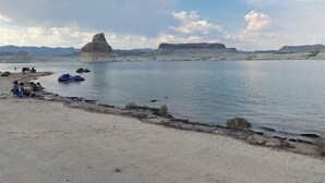 Une vue sur le lac, avec une grande formation rocheuse, des eaux calmes et une plage de sable où se trouvent des personnes et des bateaux.