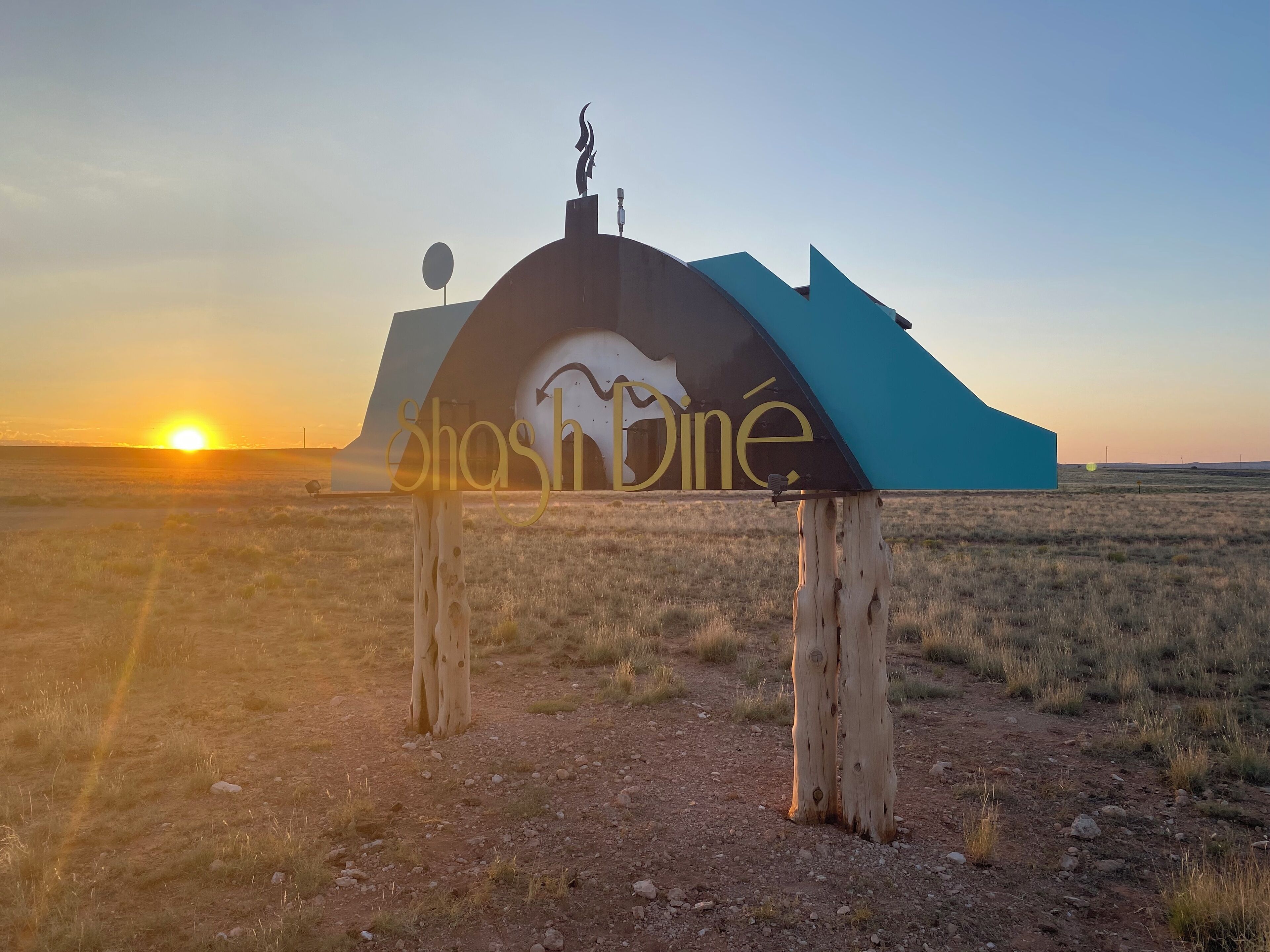 A sign with the word "Shashline" in a stylized font, mounted on a wooden post in a grassy field with a sunset in the background.