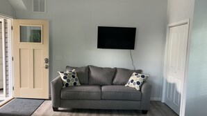 En stue med en grå sofa, to puder med mønster, et fladskærms-tv på væggen og et trægulv.