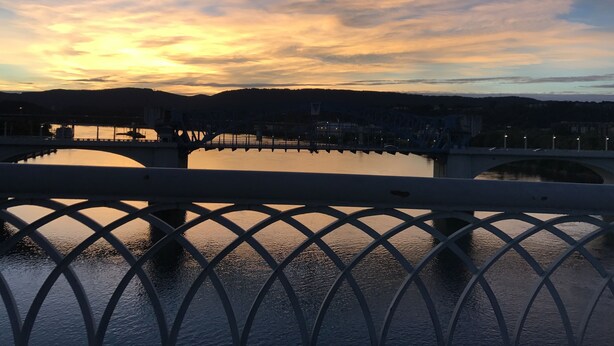 Solnedgang over en flod med en bro og et gelænder i forgrunden.