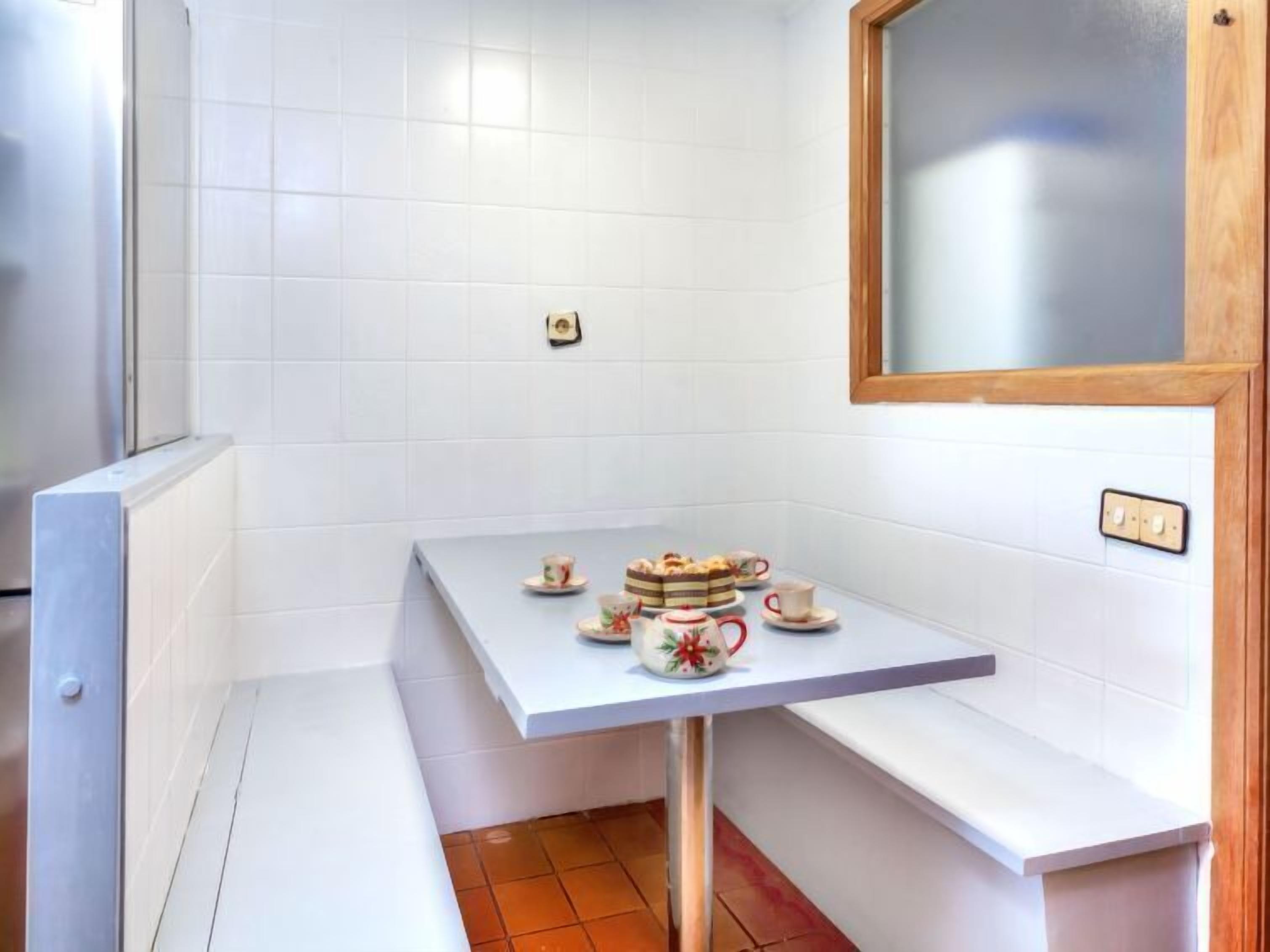 A small kitchen with white tiles, a table with tea cups and a cake, and a wooden-framed mirror.