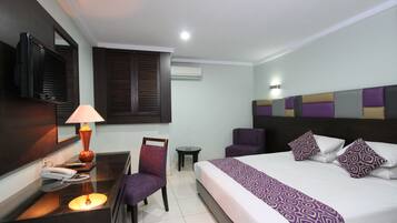 A hotel room with a bed, a desk with a lamp, a chair, a small table, and a TV.