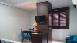 A hotel room with a bed, a desk with a chair, a TV mounted on the wall, and a window with blinds. - Hotel Alma (Jakarta)