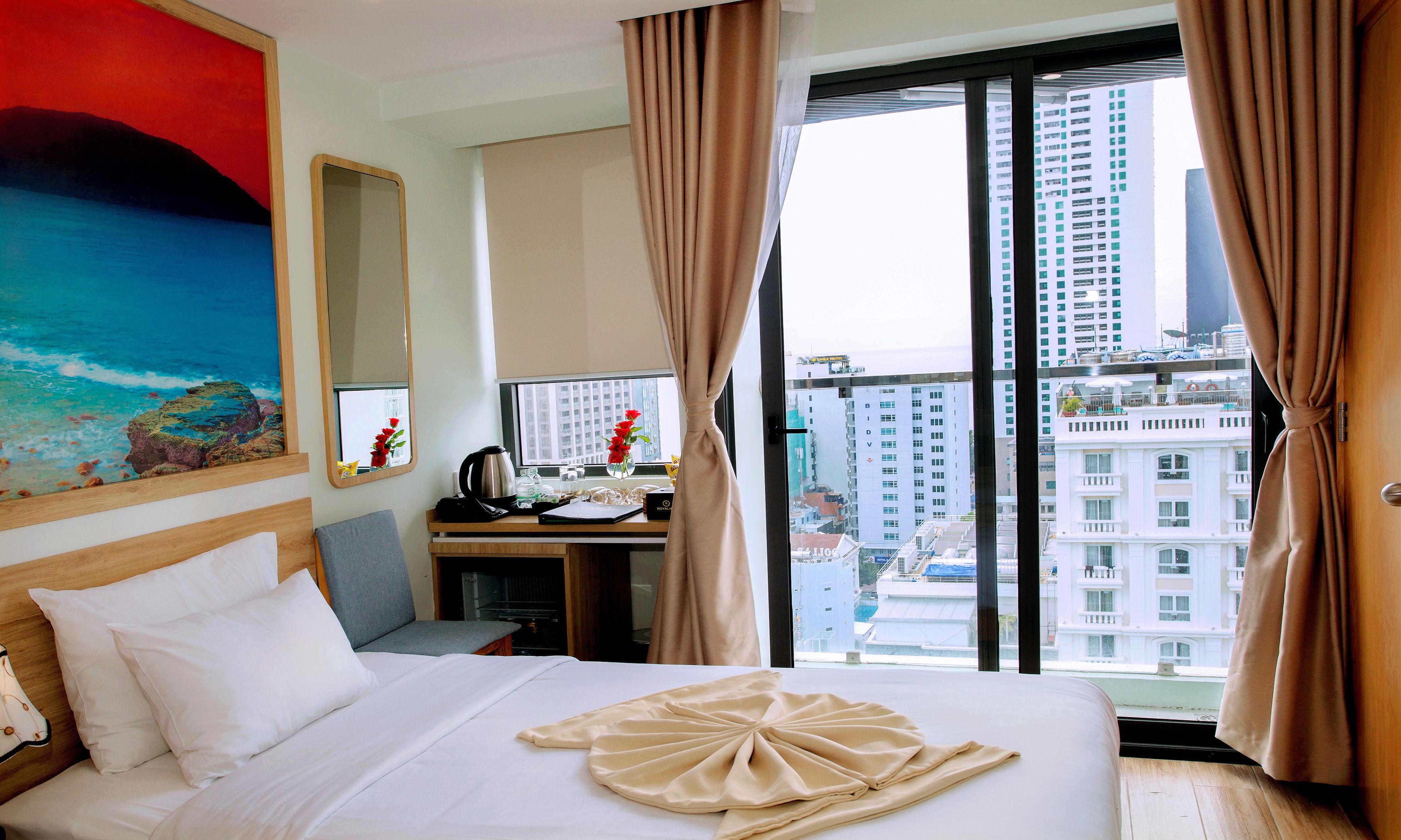 Quarto de hotel com uma cama, escrivaninha, cadeira, pintura e vista para a cidade.