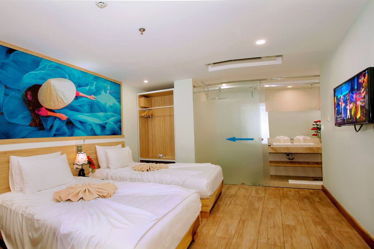A hotel room with two beds, a large painting, a wardrobe, a bathroom with a sink and mirror, and a TV on the wall.