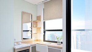 A modern, well-lit room with a desk, chair, and a large window offering a city view.