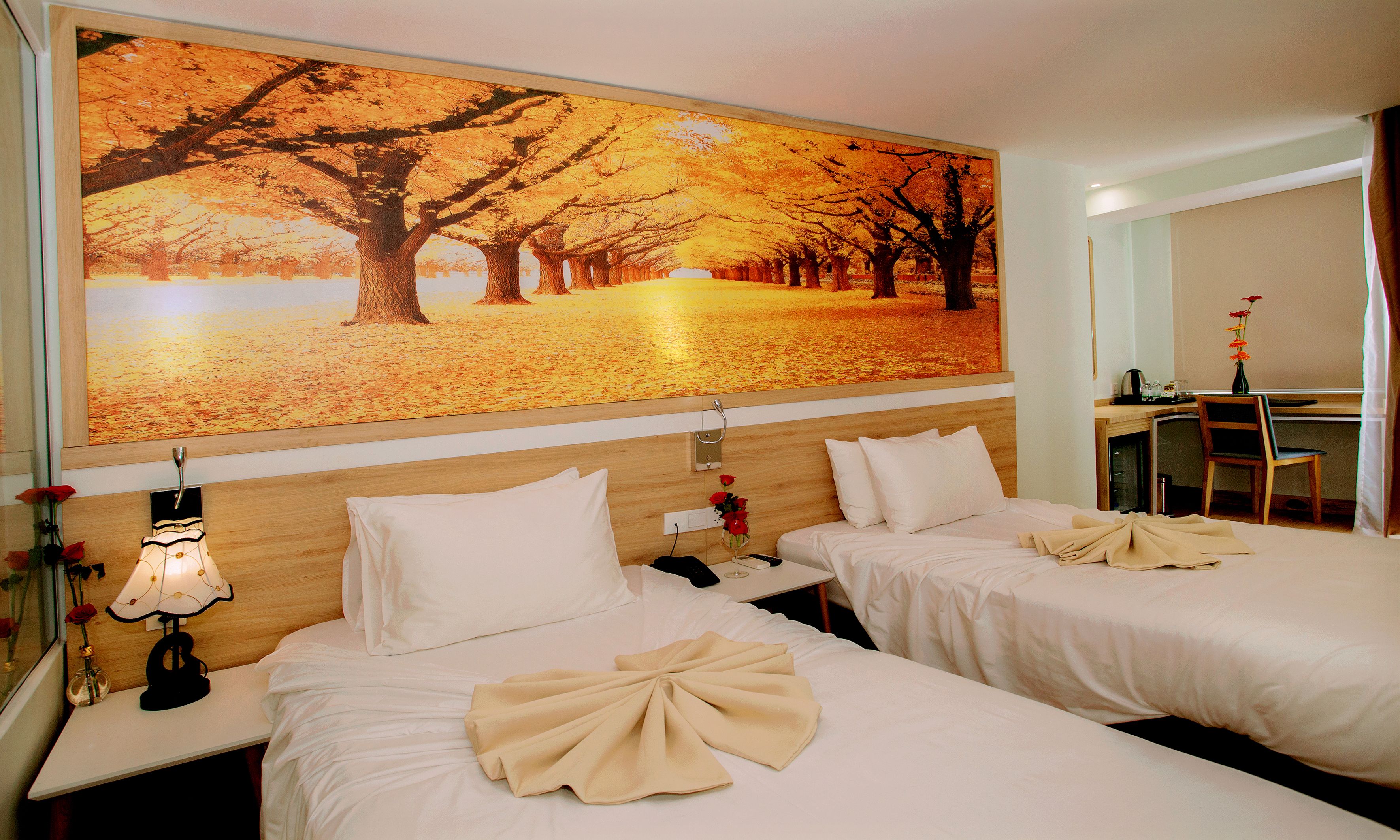 A hotel room with two beds, a desk, and a large autumn-themed painting on the wall.