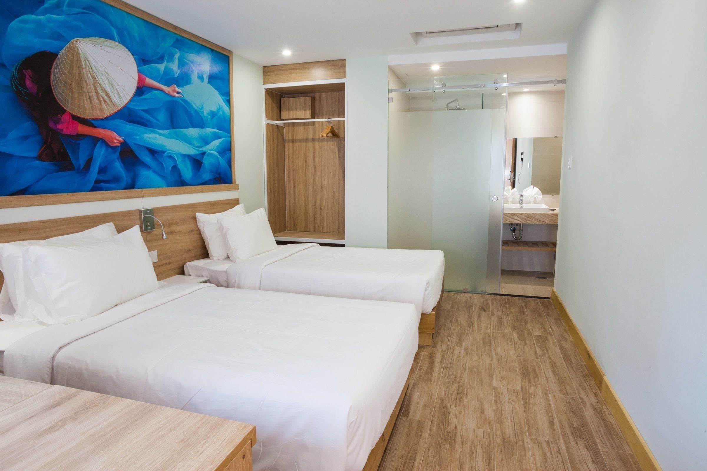 A hotel room with two beds, a wooden headboard, a large painting, a wardrobe, a bathroom with a mirror, and a wooden floor.