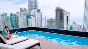 A rooftop pool with lounge chairs, overlooking a cityscape with high-rise buildings.