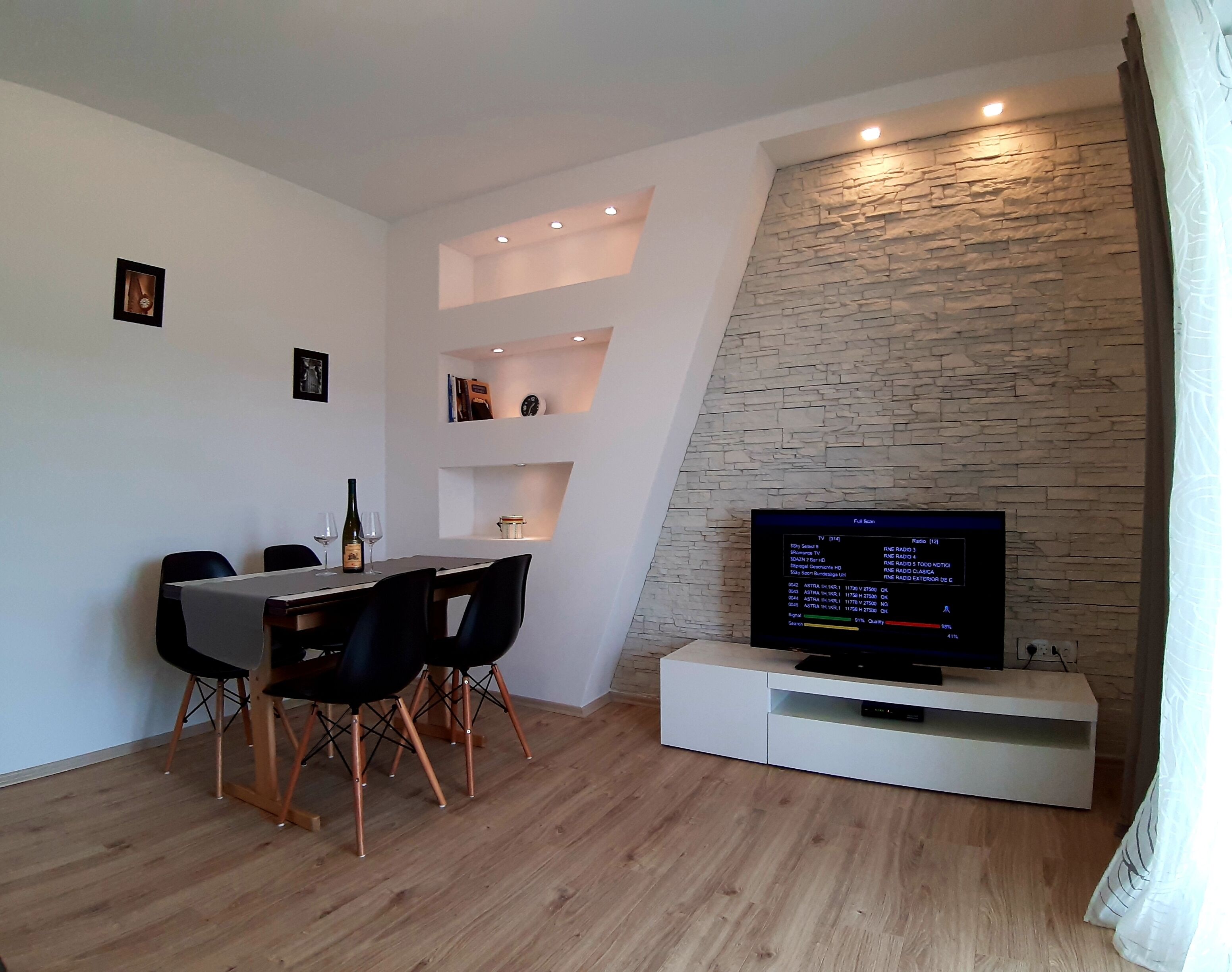 Un salon moderne comprenant un coin repas, un mur décoratif en pierre et un téléviseur à écran plat posé sur un meuble blanc.