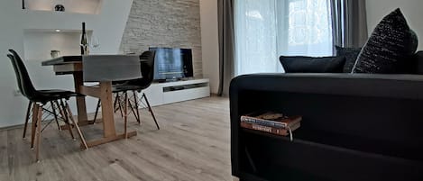 A modern living room with a black sofa, a dining table with chairs, a TV, and a bookshelf.