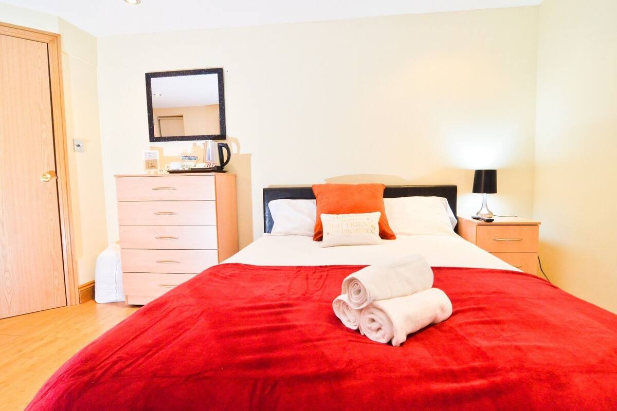 Uma cama bem arrumada com um cobertor vermelho e duas almofadas, um penteadeira de madeira, um espelho e uma mesinha de cabeceira com um abajur.