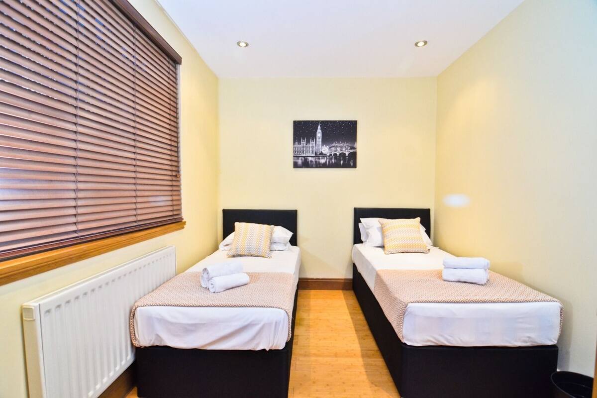 Quarto duplo com duas camas, um radiador, janela com persianas e uma fotografia da paisagem urbana na parede.