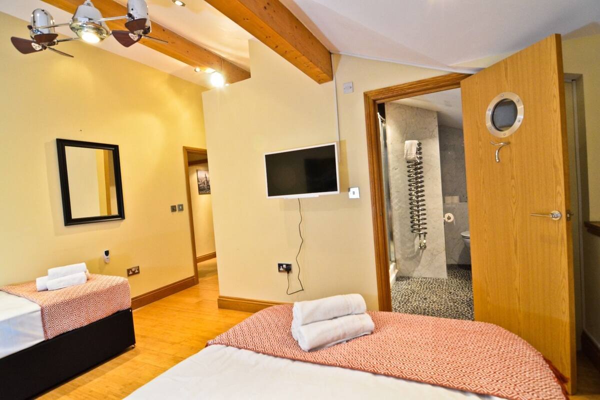 Quarto de hotel com duas camas, televisão, chuveiro e teto de madeira.