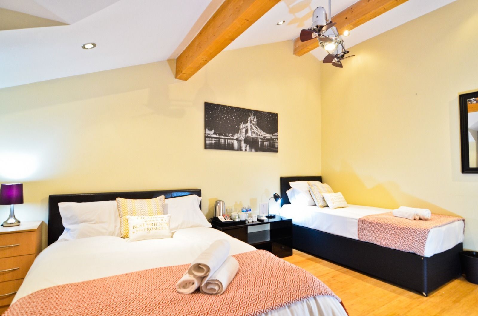 Ein Hotelzimmer mit zwei Betten, einem Nachttisch, einer Lampe, einem gerahmten Bild einer Brücke und einem Deckenventilator.
