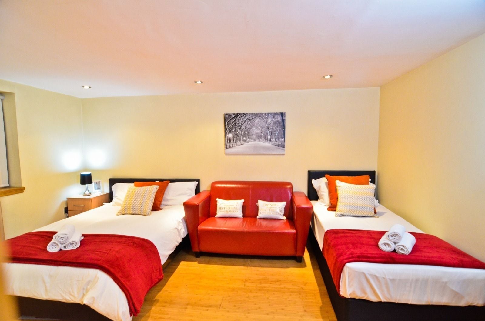 A hotel room with two single beds and a sofa bed, each with red bedding and pillows. There is a painting on the wall above the sofa bed.