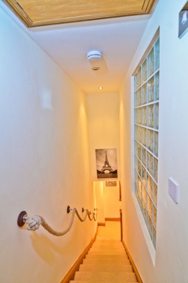 A narrow hallway with a wooden staircase, a rope pull for the door, and a glass block wall on the right.