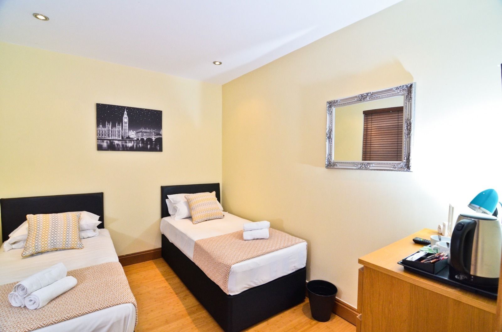Habitación de hotel con dos camas, un escritorio, un espejo, un cesto de basura y un cuadro con una vista urbana.