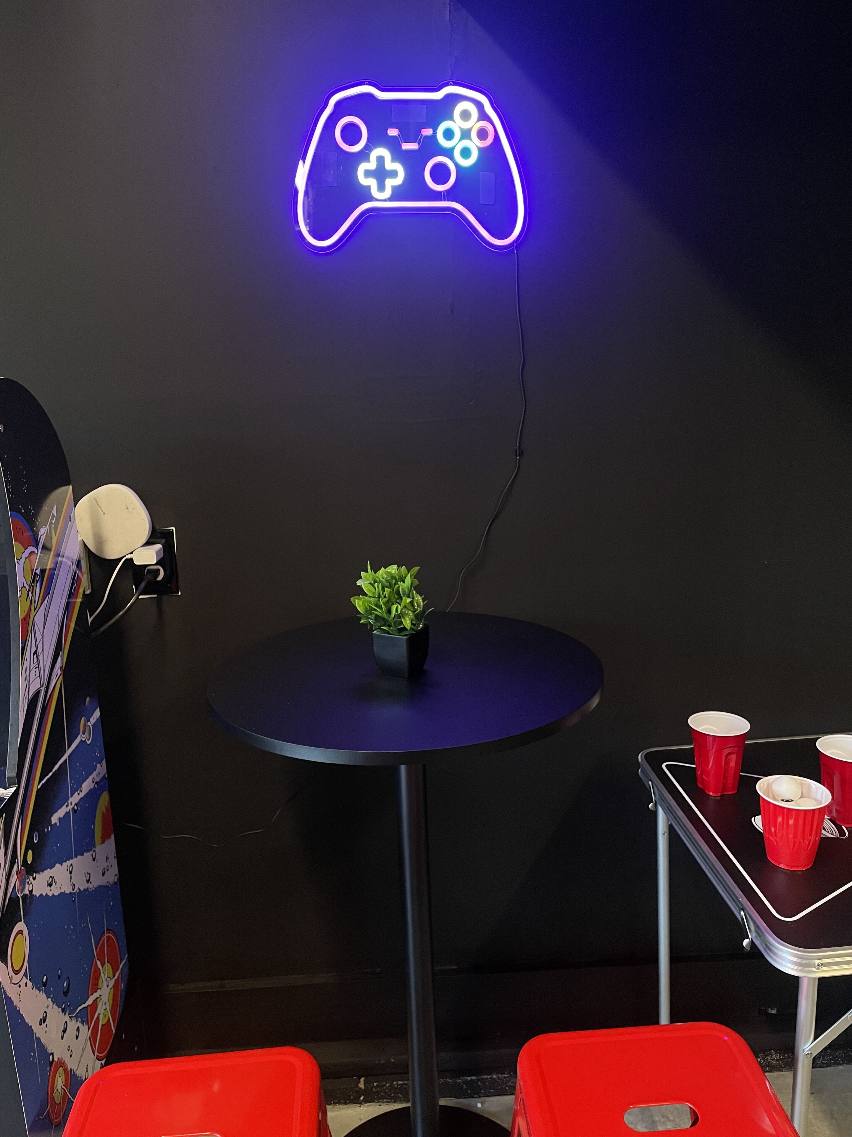 Un letrero con luces de neón que muestra un control de videojuegos, una pequeña mesa con una planta y vasos rojos sobre una bandeja.