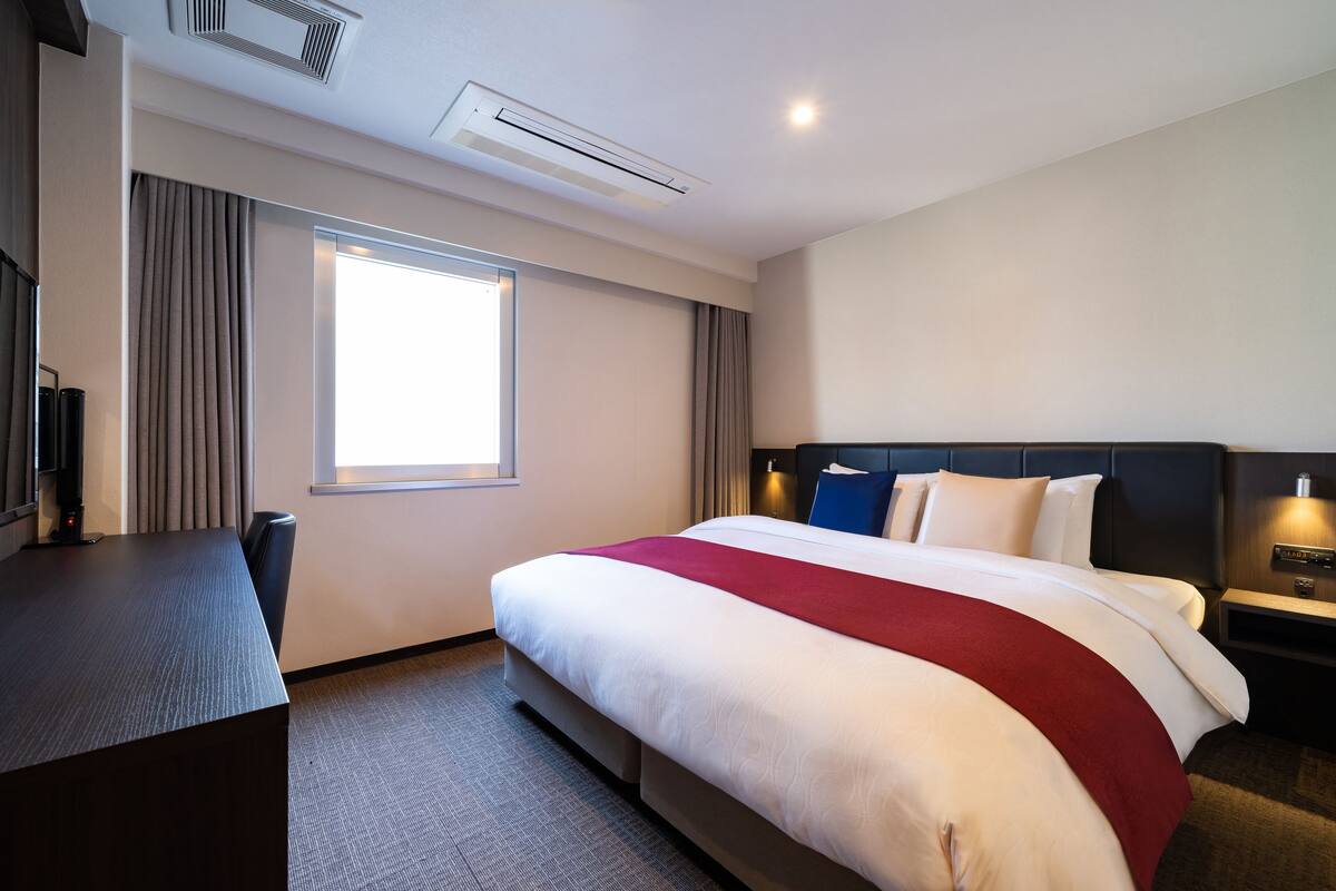 A hotel room with a large bed, a desk, a chair, a TV, and a window with curtains.