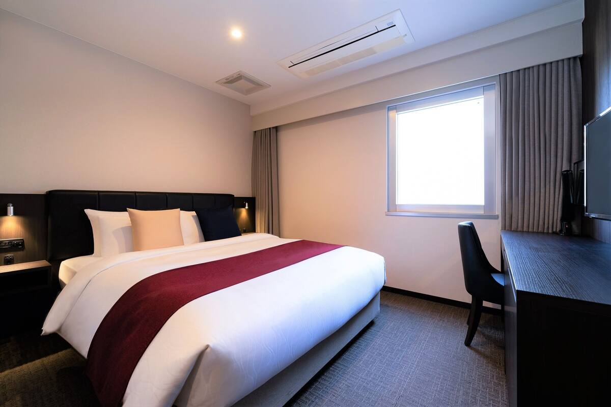 A hotel room with a large bed, a desk, a chair, and a window with curtains.
