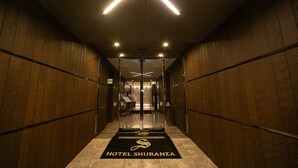 Een hotelgang met houten lambrisering, een zwart tapijt met het logo 'HOTEL SHURANZA' en een glazen lift.