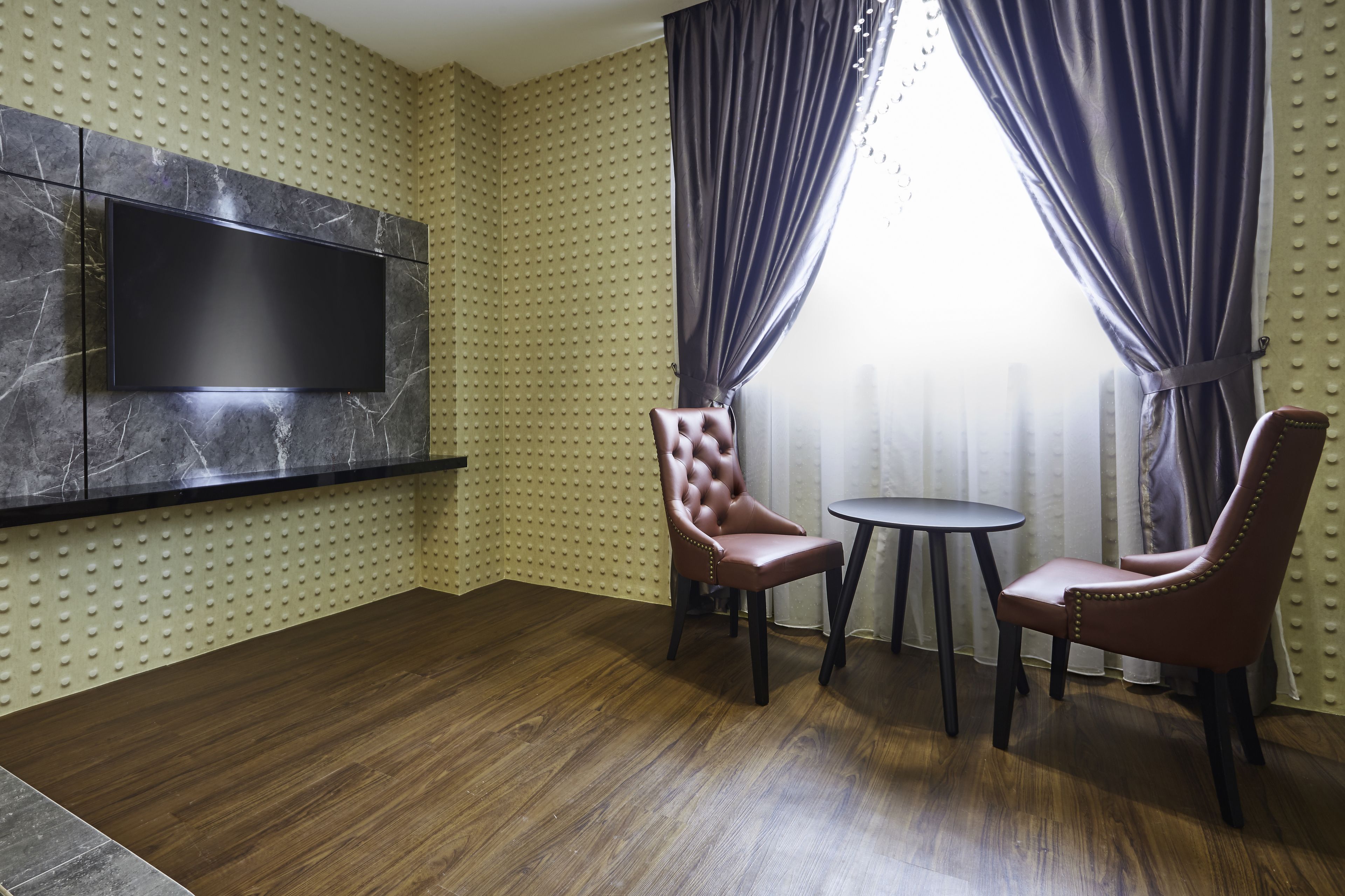 A room with a flat-screen TV mounted on a marble wall, two leather chairs around a small table, and a window with curtains.