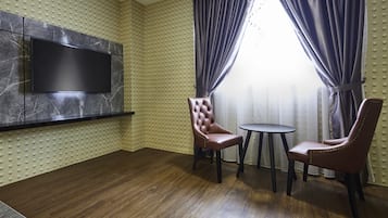 A room with a flat-screen TV mounted on a marble wall, two leather chairs around a small table, and a window with curtains.