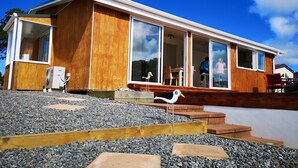 Une maison moderne dotée de grandes fenêtres, d’un revêtement extérieur en bois et d’un petit espace extérieur aménagé pour s’asseoir.