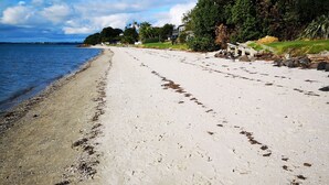 Une plage de sable parsemée d’empreintes, un banc en bois et, en arrière-plan, des arbres et des maisons.