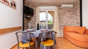 Zona pranzo con tavolo apparecchiato per quattro persone, una poltrona e un televisore montato a parete, e una parete con texture in pietra.