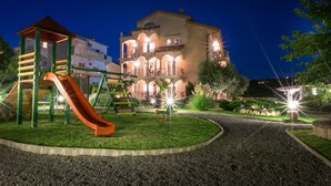 A playground with a slide and climbing structure, surrounded by a well-lit garden area with a building in the background.