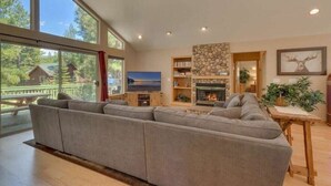 A spacious living room with a large gray sectional sofa, a stone fireplace, a television, and a view of a lake through large windows.