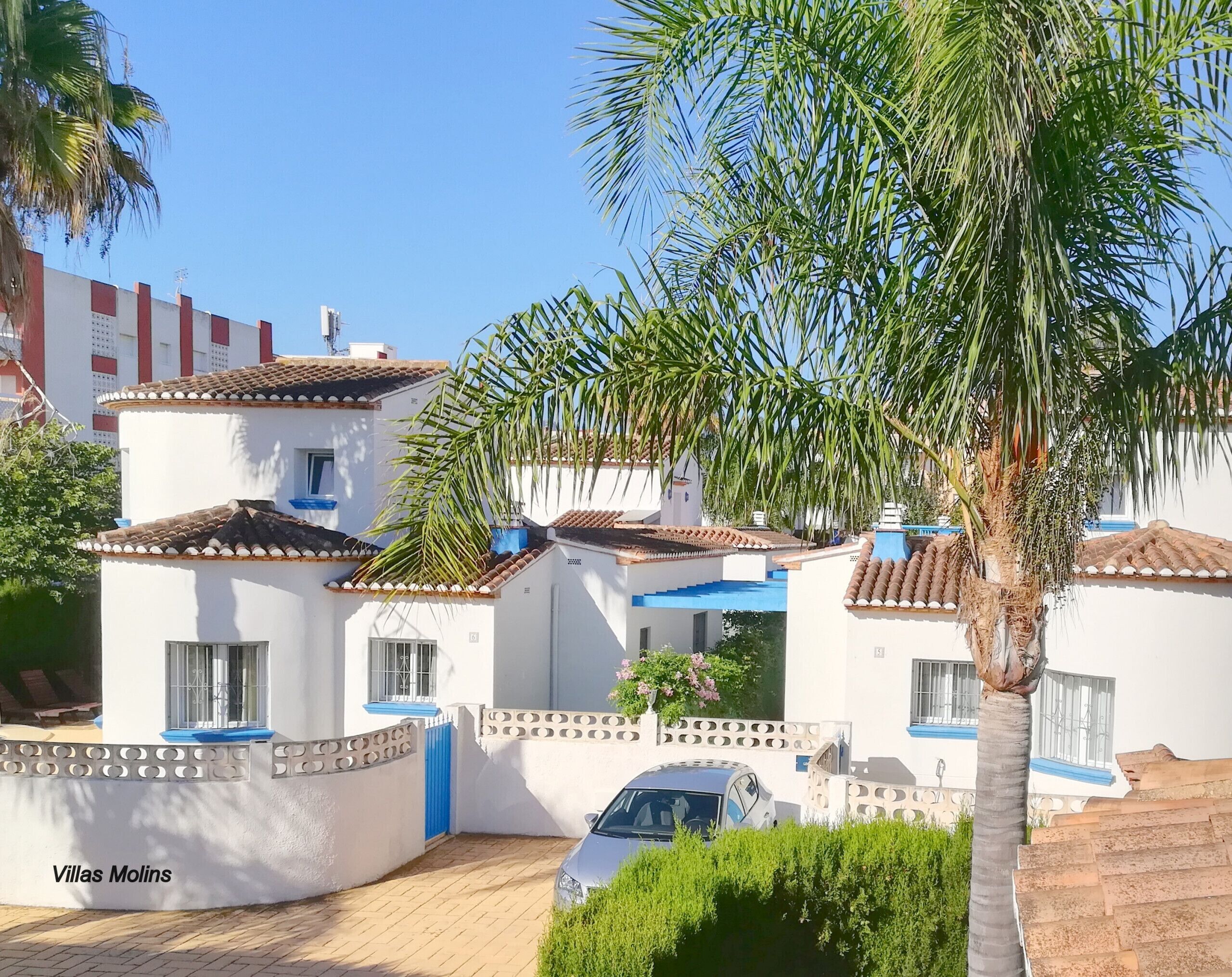 Un conjunto de villas blancas con detalles azules, un coche estacionado en la entrada y una palmera en primer plano.