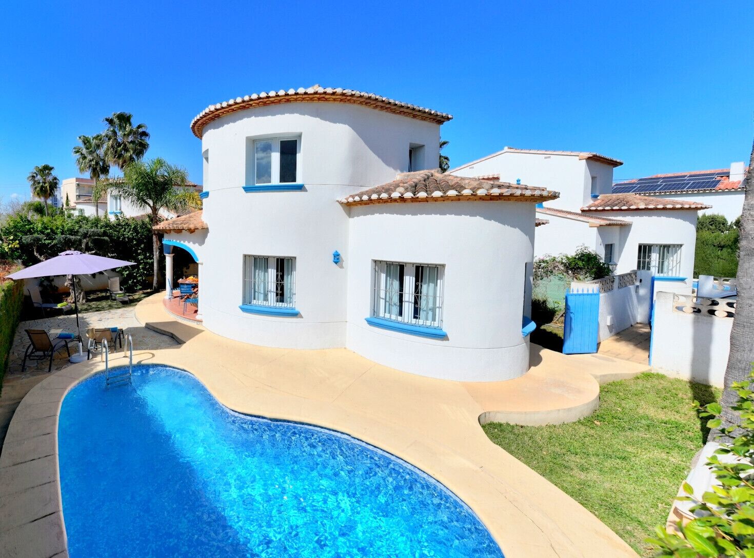 A two-story house with a curved swimming pool, a patio with chairs, and a small garden area.