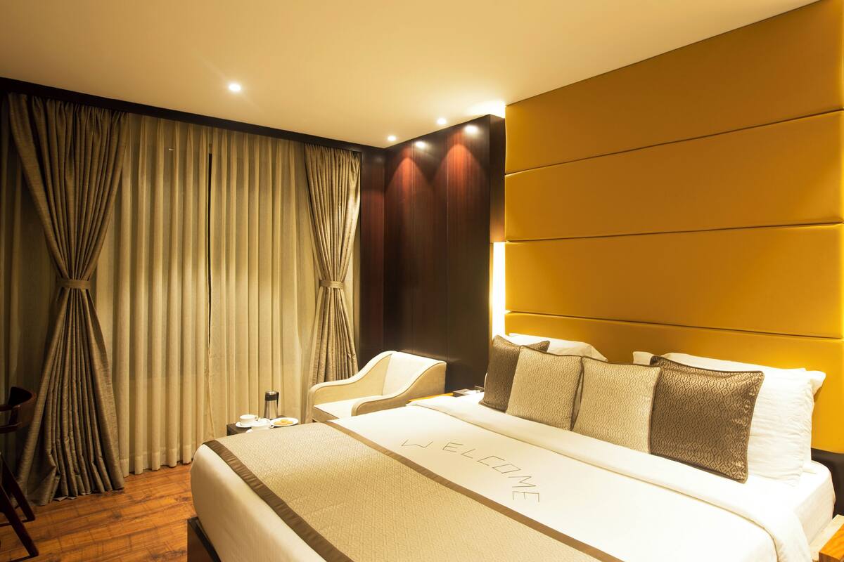 A modern hotel room with a large bed, a headboard with built-in lighting, a seating area with a chair, and a large window with curtains.