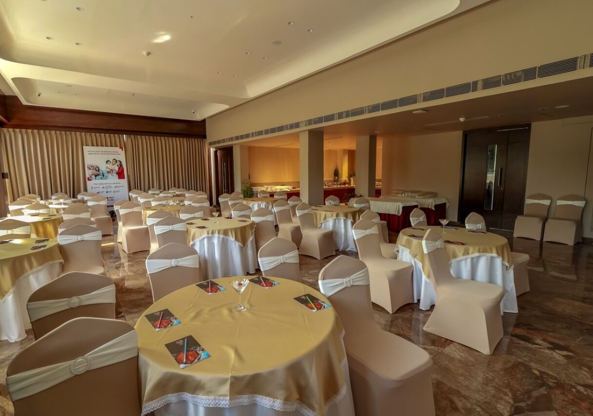 Salão de banquetes com mesas redondas cobertas por toalhas brancas e cadeiras com capas brancas.
