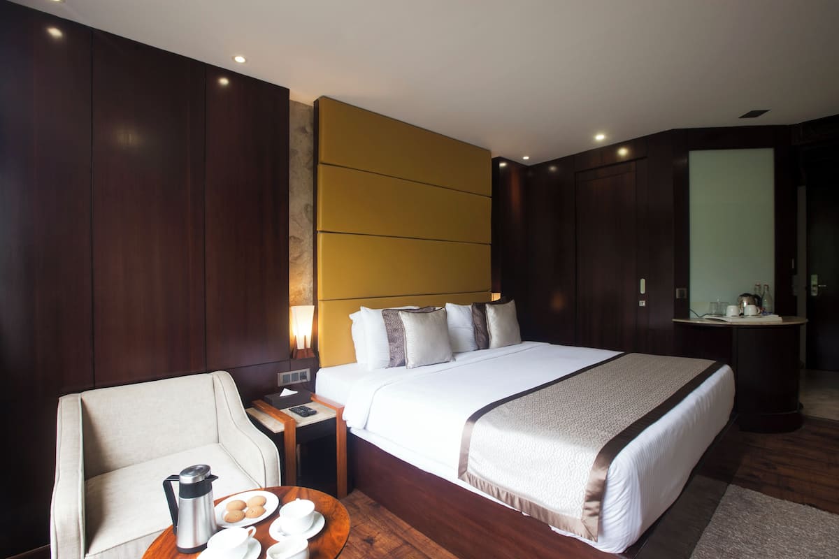 A hotel room with a large bed, a bedside table with a lamp, a chair, and a coffee table with cups and a kettle.