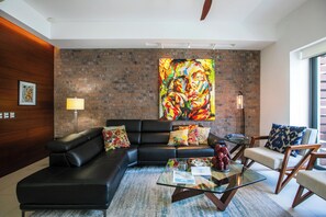 Un salon moderne avec un canapé en cuir noir, une table basse en verre et un tableau abstrait aux couleurs vives accroché au mur de briques.