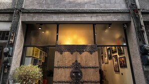 Entrada de un edificio con una puerta de madera grande, una planta en maceta y una escalera que sube a los pisos superiores.