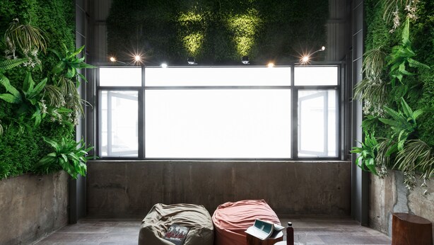 Una habitación con un ventanal grande, plantas verdes, dos pufs y una pequeña mesa de madera.