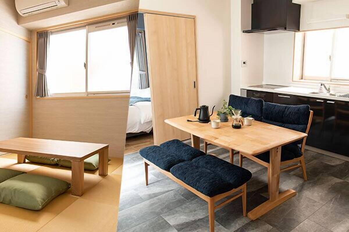 Una habitación con una mesa de madera, bancos, una cama, un ventanal y una zona de cocina.