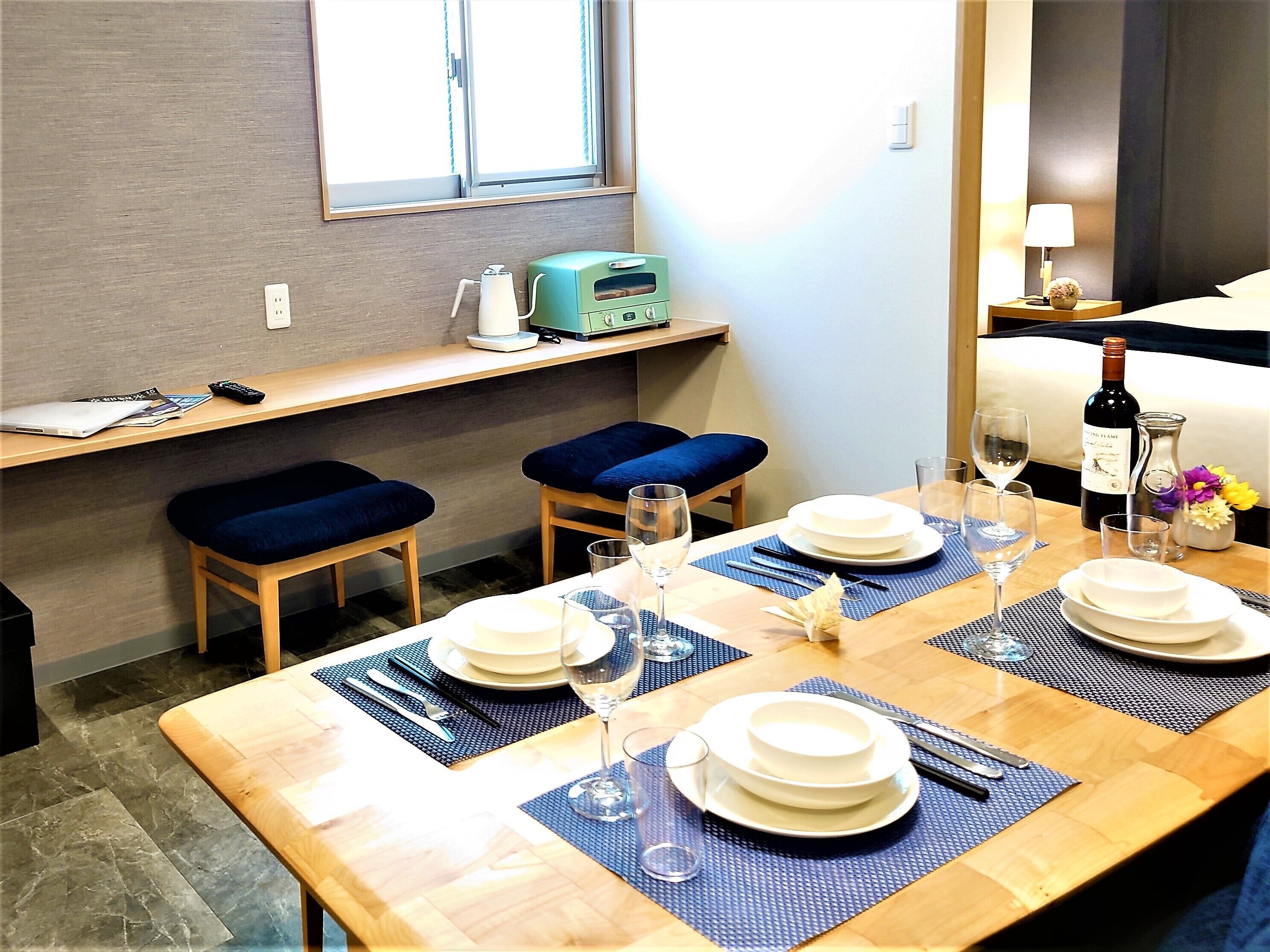 A dining area with a wooden table set for four, blue cushioned chairs, a small kitchen area with a microwave, and a window with curtains.