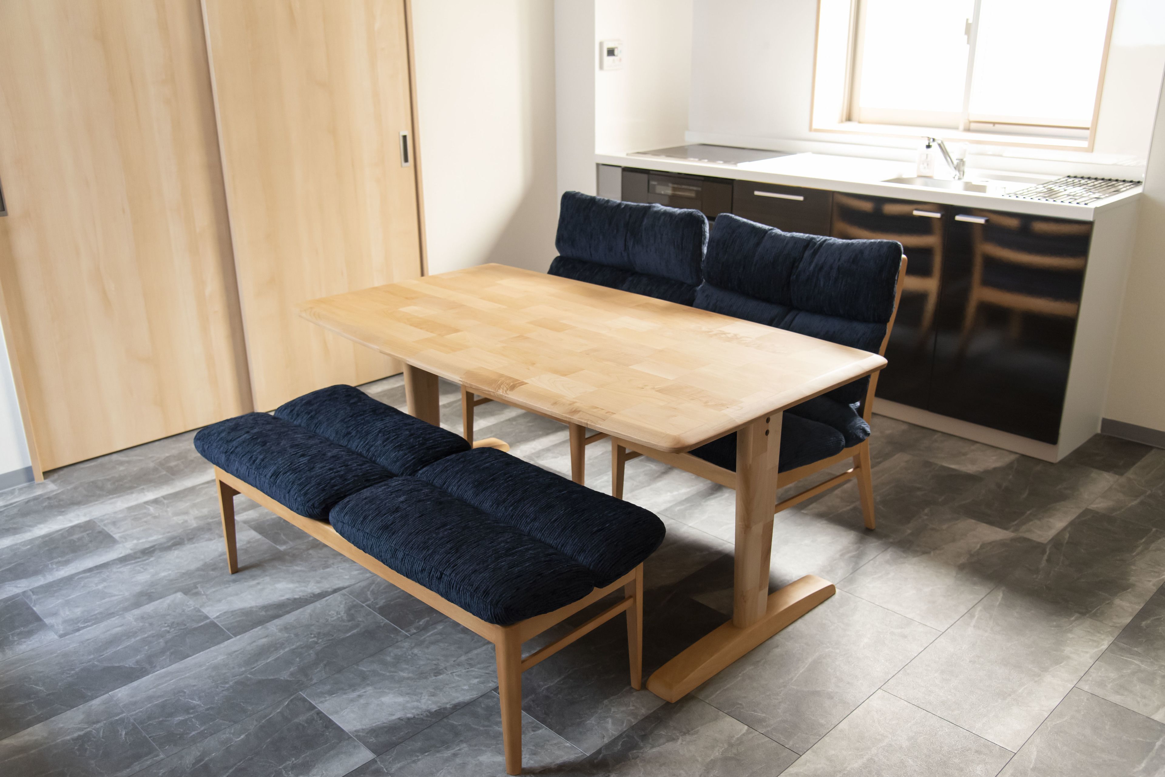 Een compacte keuken met een houten tafel en bankje, vloer van grijze tegels, en een raam dat natuurlijk licht binnenlaat.