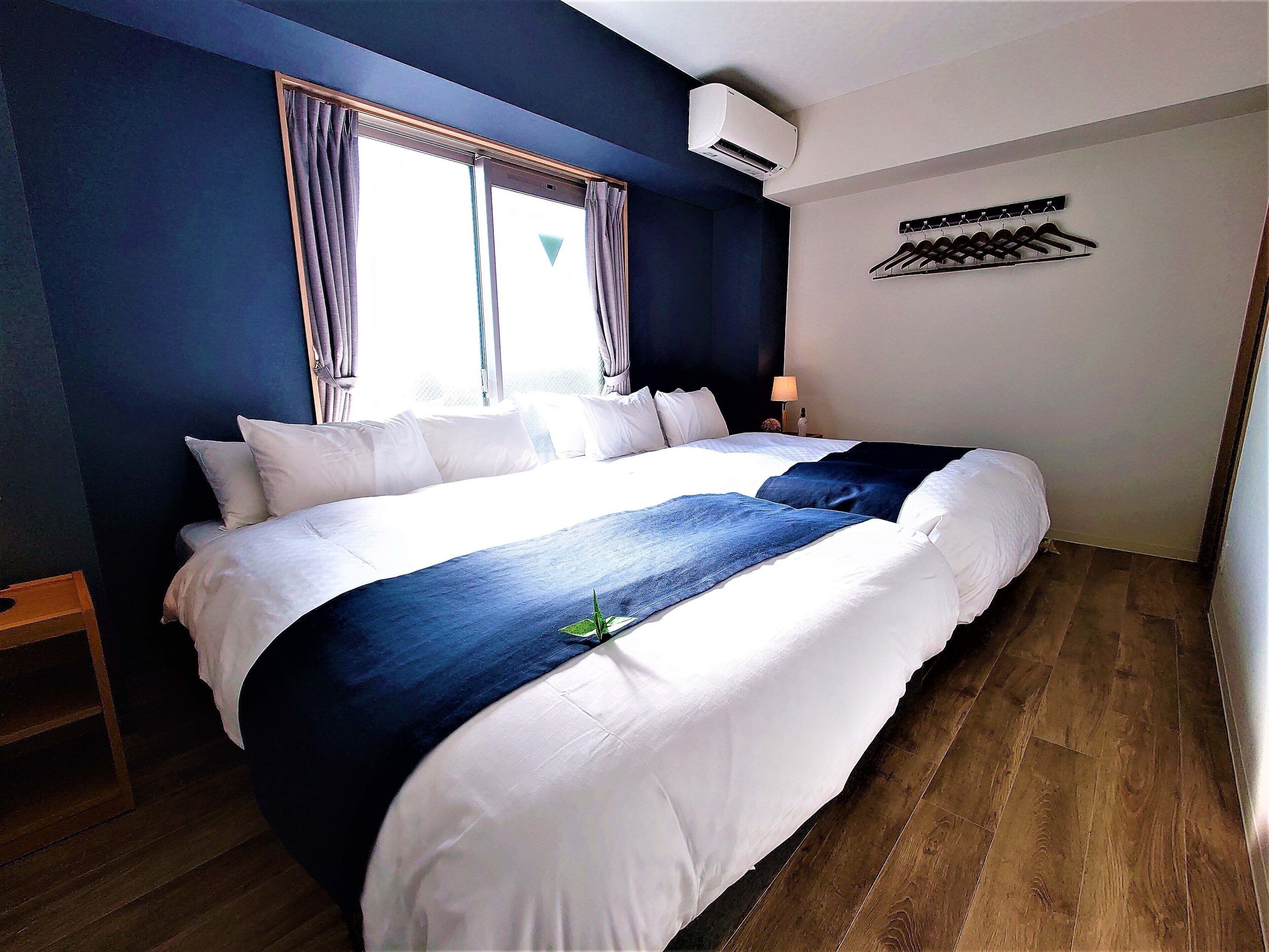 Una cama bien hecha con ropa de cama blanca y azul, una ventana con cortinas, un aire acondicionado de pared y un perchero con ganchos.